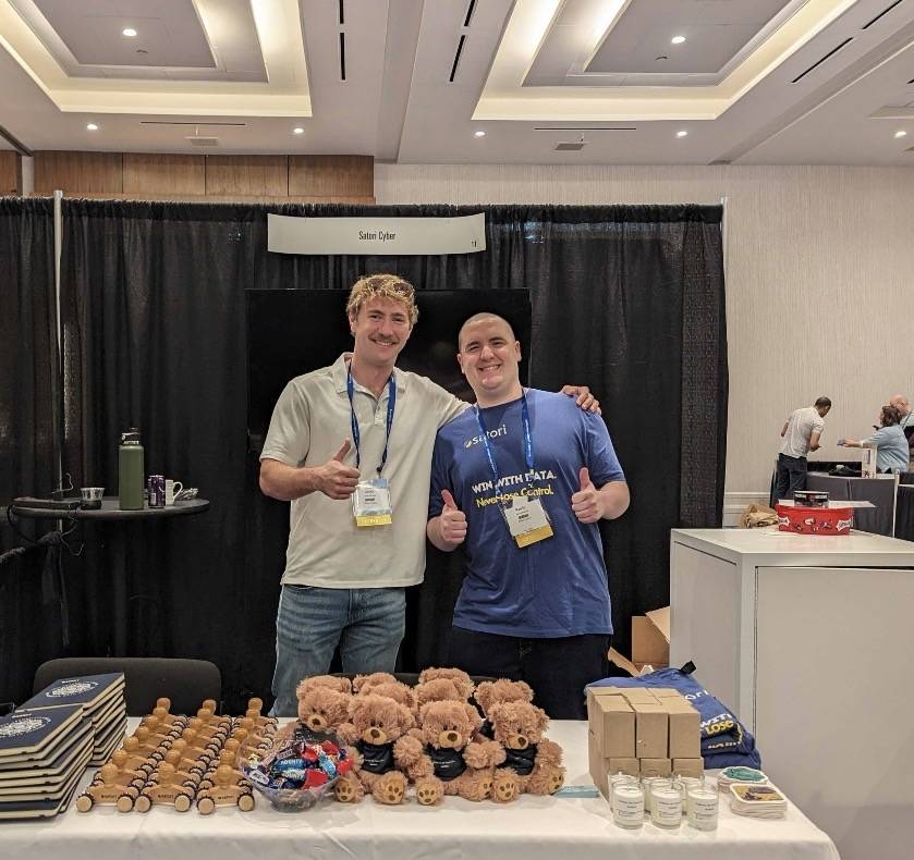 Two Richey Consulting team members giving thumbs-up behind a conference booth table with branded teddy bears and notebooks.